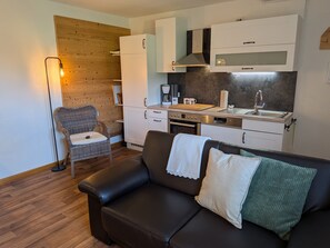 Et moderne køkken med hvide skabe, indbygget ovn og vask. Der er en fletstol og en sort lædersofa med grøn pude.