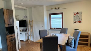 Zona pranzo con tavolo e sedie in legno, cucina con pensili bianchi, una scala di legno e una finestra con persiane.