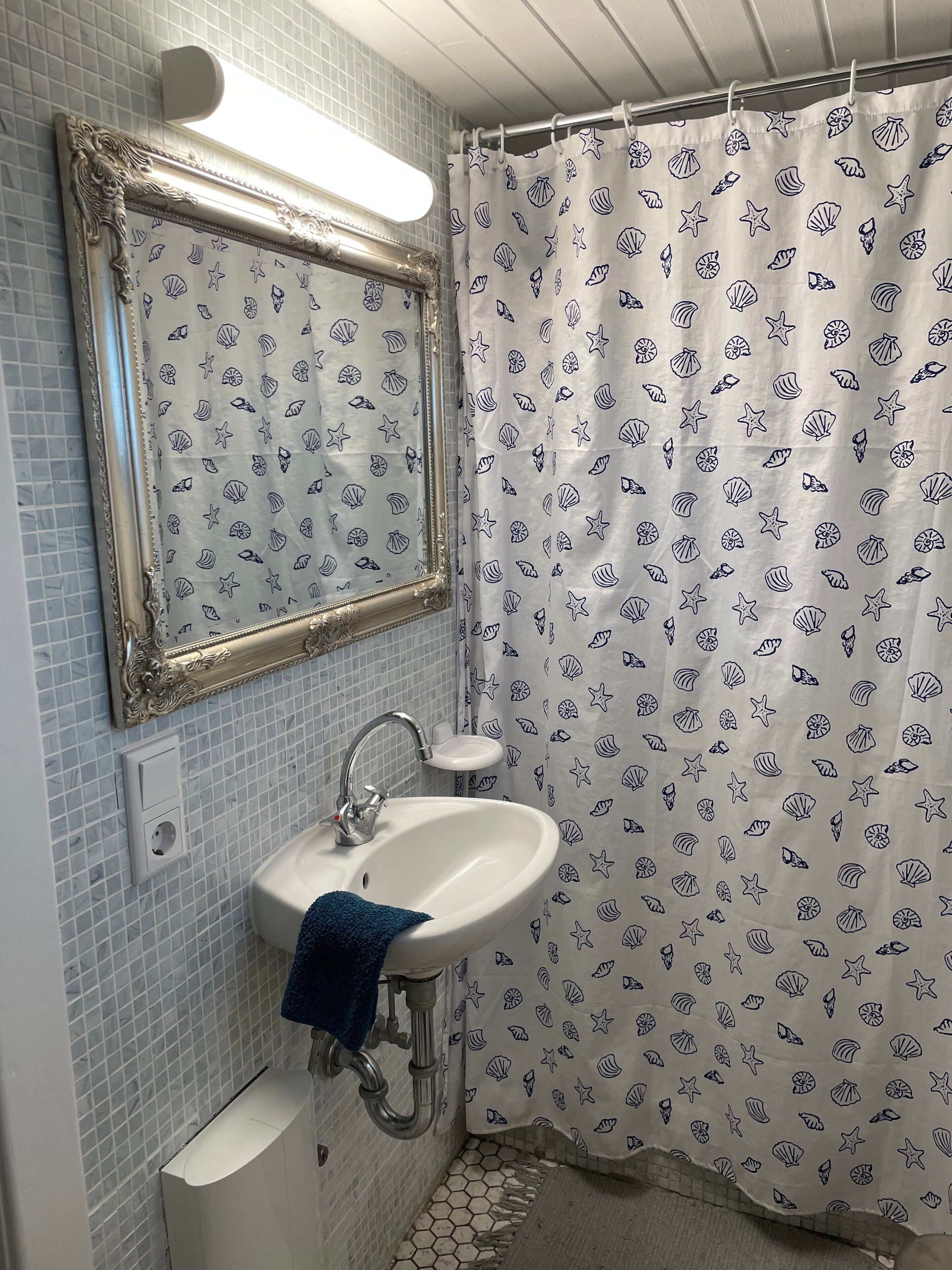 A bathroom with a tiled wall, a sink with a faucet, a mirror with an ornate frame, and a shower curtain with a sea shell pattern.