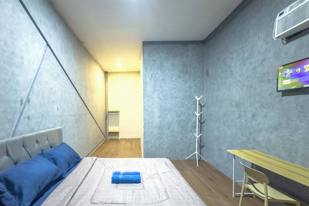A bedroom with a bed, a desk, a chair, a TV, and a coat rack.