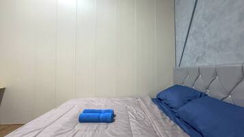 A neatly made bed with a grey bedspread and blue pillows, a white wall with vertical paneling, and a window with a grey blind.