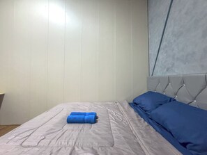 A neatly made bed with a grey bedspread and blue pillows, a white wall with vertical paneling, and a window with a grey blind.