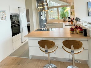 A modern kitchen with a central island, two bar stools, a refrigerator, and a window with a view of greenery. - Stunning sunrises over Harre Vig (Roslev)
