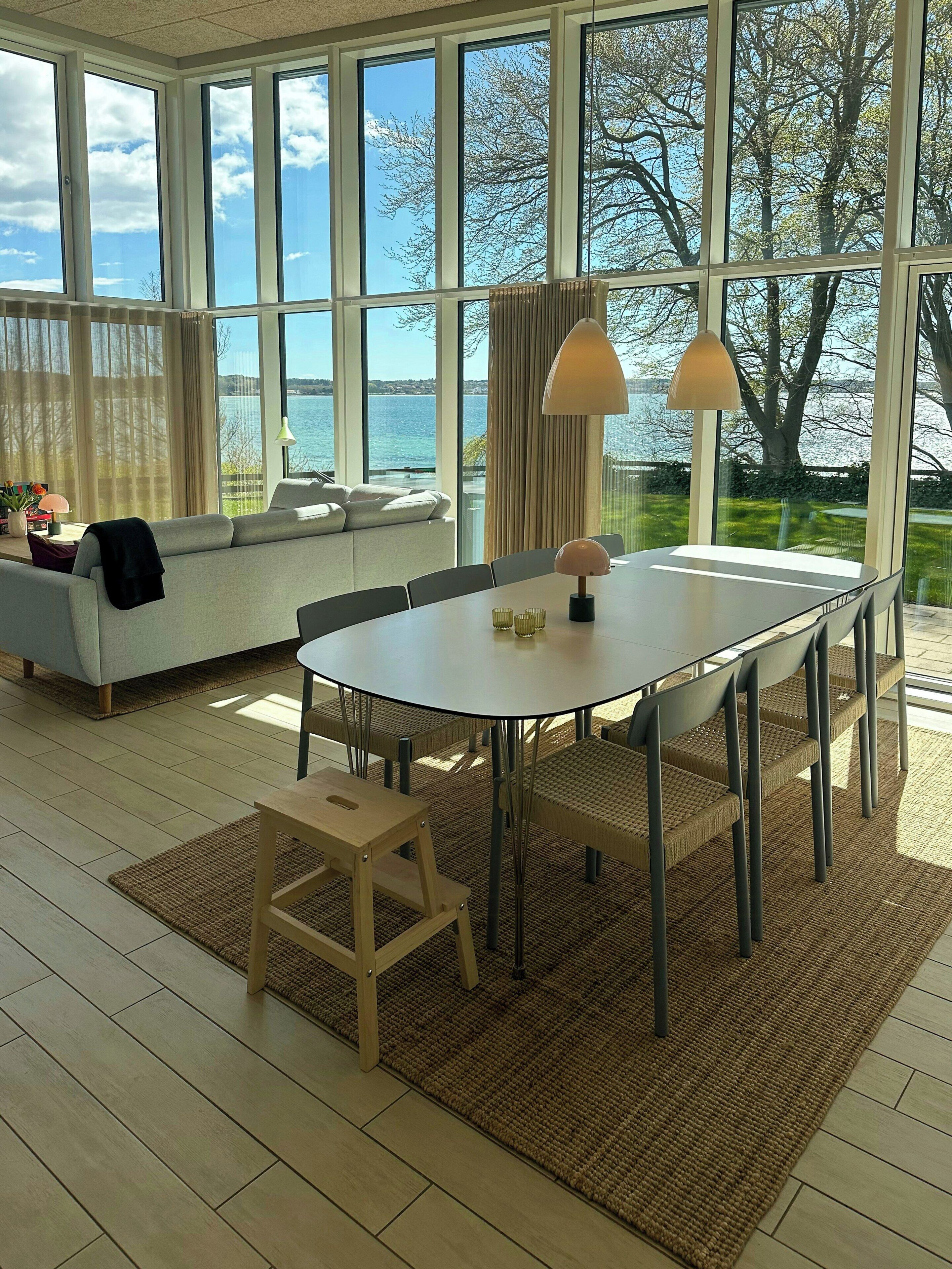 A modern living room with a large window, a sofa, a dining table, and a view of the sea.