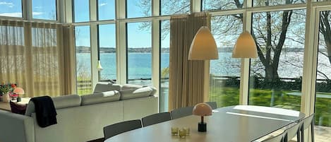 A modern living room with a large window, a sofa, a dining table, and a view of the sea.