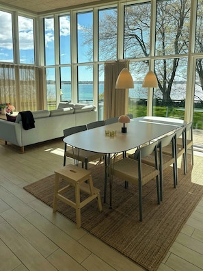 Ein modernes Wohnzimmer mit großem Fenster, einer Couch, einem Esstisch und Blick aufs Meer.
