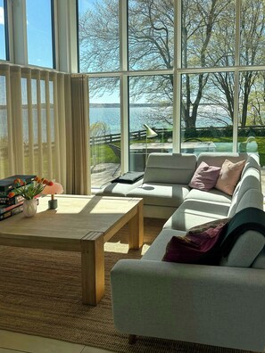 Ein modernes Wohnzimmer mit großem Fenster, einer Couch, einem Couchtisch und Blick auf ein Gewässer.