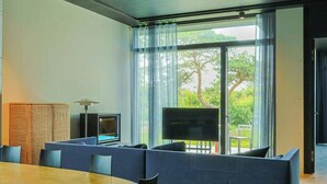 Ein modernes Wohnzimmer mit großem Fernseher, einer Sitzecke und Blick auf Grün durch große Fenster.