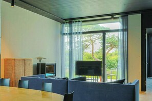 Un salon moderne doté d’un grand téléviseur, d’un espace salon et offrant une vue sur la verdure grâce à de grandes fenêtres.