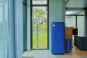 Una habitación moderna con un gran ventanal corredero, un refrigerador azul, un cesto de basura y una silla de mimbre.