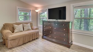 Una habitación con sofá color beige, cómoda de madera oscura, televisor de pantalla plana y ventanas amplias con vista a árboles.