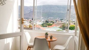 Un balcón con mesa y sillas, vista a un pueblo y montañas, y una ventana con cortinas.