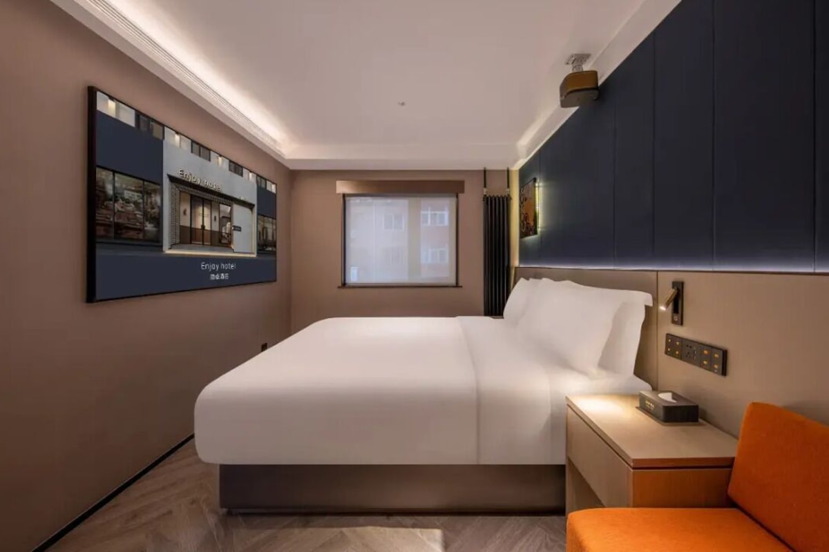 Une chambre d’hôtel moderne avec un grand lit, une table de chevet avec une lampe, une petite chaise orange, une fenêtre avec des stores et un téléviseur fixé au mur qui affiche le logo de l’hôtel.