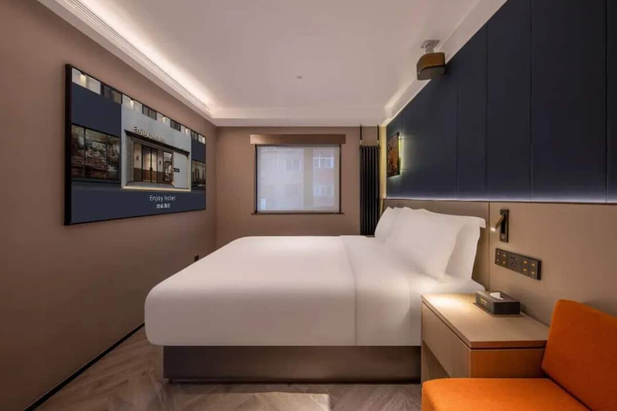 A modern hotel room with a large bed, a bedside table with a lamp, a small orange chair, a window with blinds, and a wall-mounted TV displaying a hotel sign.