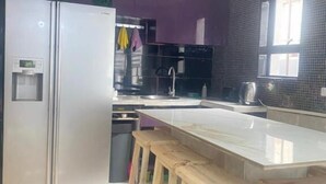 A modern kitchen with a stainless steel refrigerator, a marble countertop, wooden stools, and purple cabinets. - Quaaamiii Accomodation (Cape Town)
