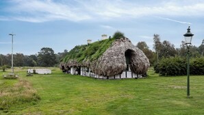 Uma construção com telhado de palha, rodeada por um campo gramado e árvores.