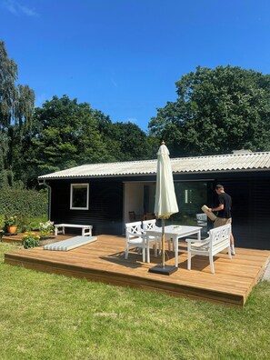 Een terras met een tafel en stoelen, een witte parasol, en een persoon met een tas.