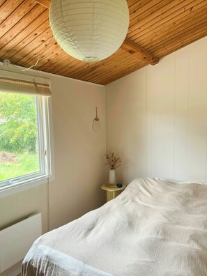 A bedroom with a wooden ceiling, a large bed, a window with a view of greenery, and a bedside table with a vase. - Summer house near Ahl Beach and Ebeltoft (Ebeltoft)