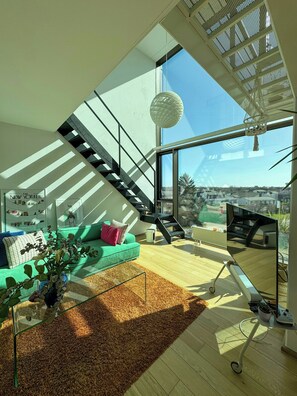 Una sala de estar moderna con un sofá verde, una escalera, un ventanal y vistas de la ciudad.