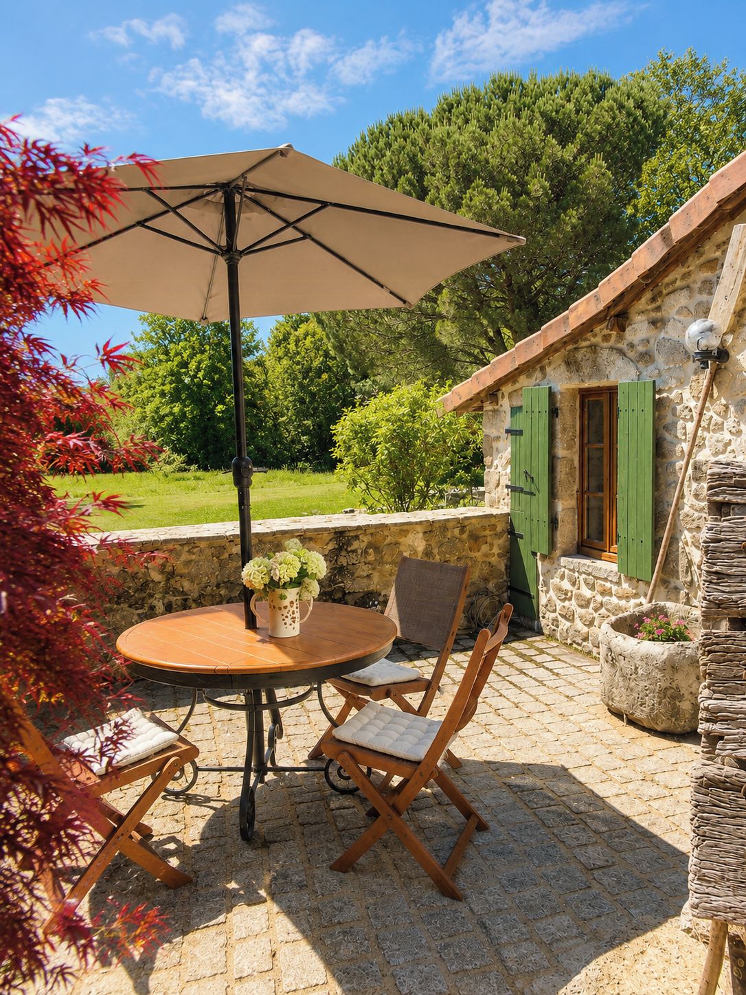 An outdoor patio with a round table, two chairs, a beige umbrella, and a stone wall.