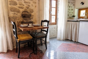 A rustic kitchen with a stone wall, a wooden table with chairs, a sink, and a window with curtains. - Cottage in former bakery in Dordogne (Saint-Estèphe)