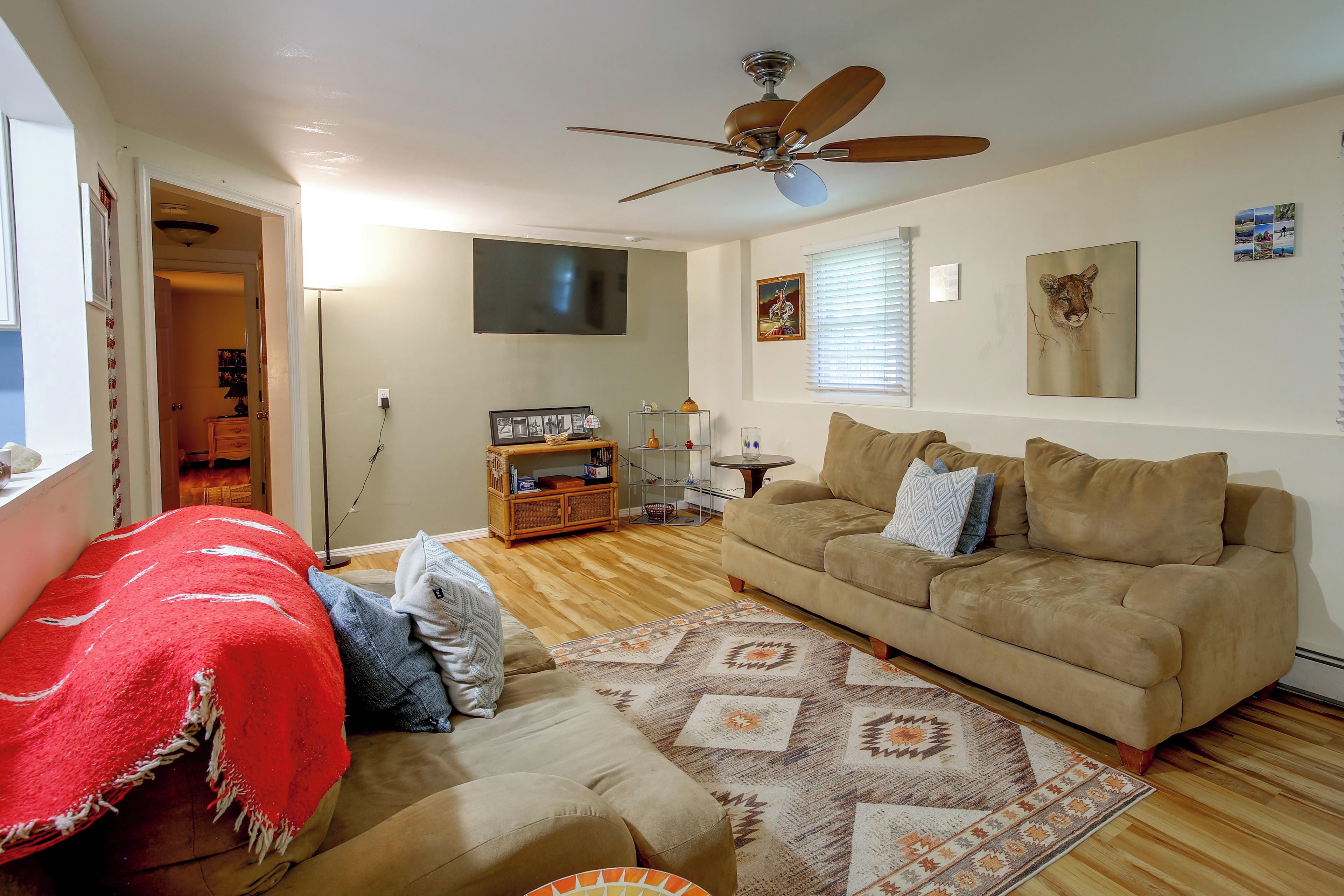 Un salon avec un ventilateur de plafond, un canapé, une couverture rouge, un tapis à motifs et un téléviseur fixé au mur.
