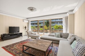 Ein modernes Wohnzimmer mit Fernseher, einer Couch, einem Couchtisch und einem großen Fenster mit Blick auf ein Gebäude und Grünflächen.