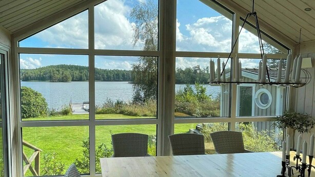 A wooden table with wicker chairs, large windows offering a view of a lake and greenery, and a hanging light fixture. - Lake cottage with private pier at Lake Nömmen (Stensjön)