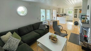 A modern living room with a dark green sofa, a white coffee table, and a grey armchair. A kitchen is visible in the background with white cabinets and a fireplace. - Lake cottage with private pier at Lake Nömmen (Stensjön)