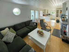 A modern living room with a dark green sofa, a white coffee table, and a grey armchair. A kitchen is visible in the background with white cabinets and a fireplace. - Lake cottage with private pier at Lake Nömmen (Stensjön)