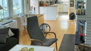 A modern living room with a sofa, a dining table with a bowl and candles, a fireplace, and a kitchen in the background. - Lake cottage with private pier at Lake Nömmen (Stensjön)