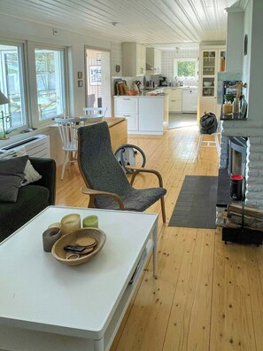 A modern living room with a sofa, a dining table with a bowl and candles, a fireplace, and a kitchen in the background. - Lake cottage with private pier at Lake Nömmen (Stensjön)