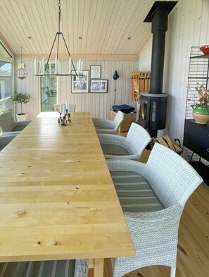 A long wooden dining table with wicker chairs, a hanging light fixture, a black stove, and framed pictures on the wall. - Lake cottage with private pier at Lake Nömmen (Stensjön)