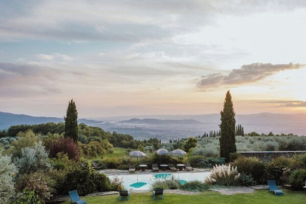 Ein Pool mit Liegestühlen und Sonnenschirmen, umgeben von viel Grün und Blick auf ferne Berge.