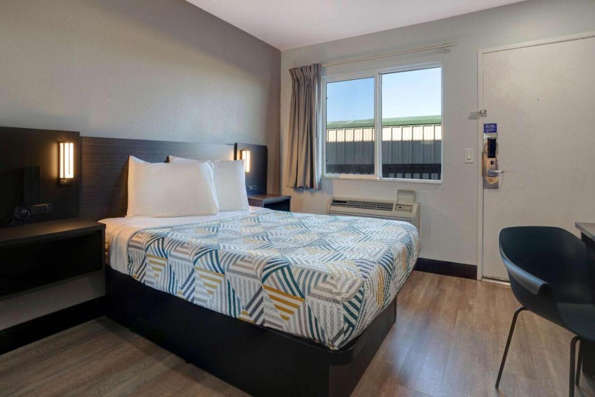 A hotel room with a bed, a desk, a chair, a TV, a window with curtains, and an air conditioning unit.