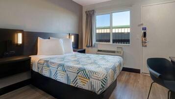 A hotel room with a bed, a desk, a chair, a TV, a window with curtains, and an air conditioning unit.