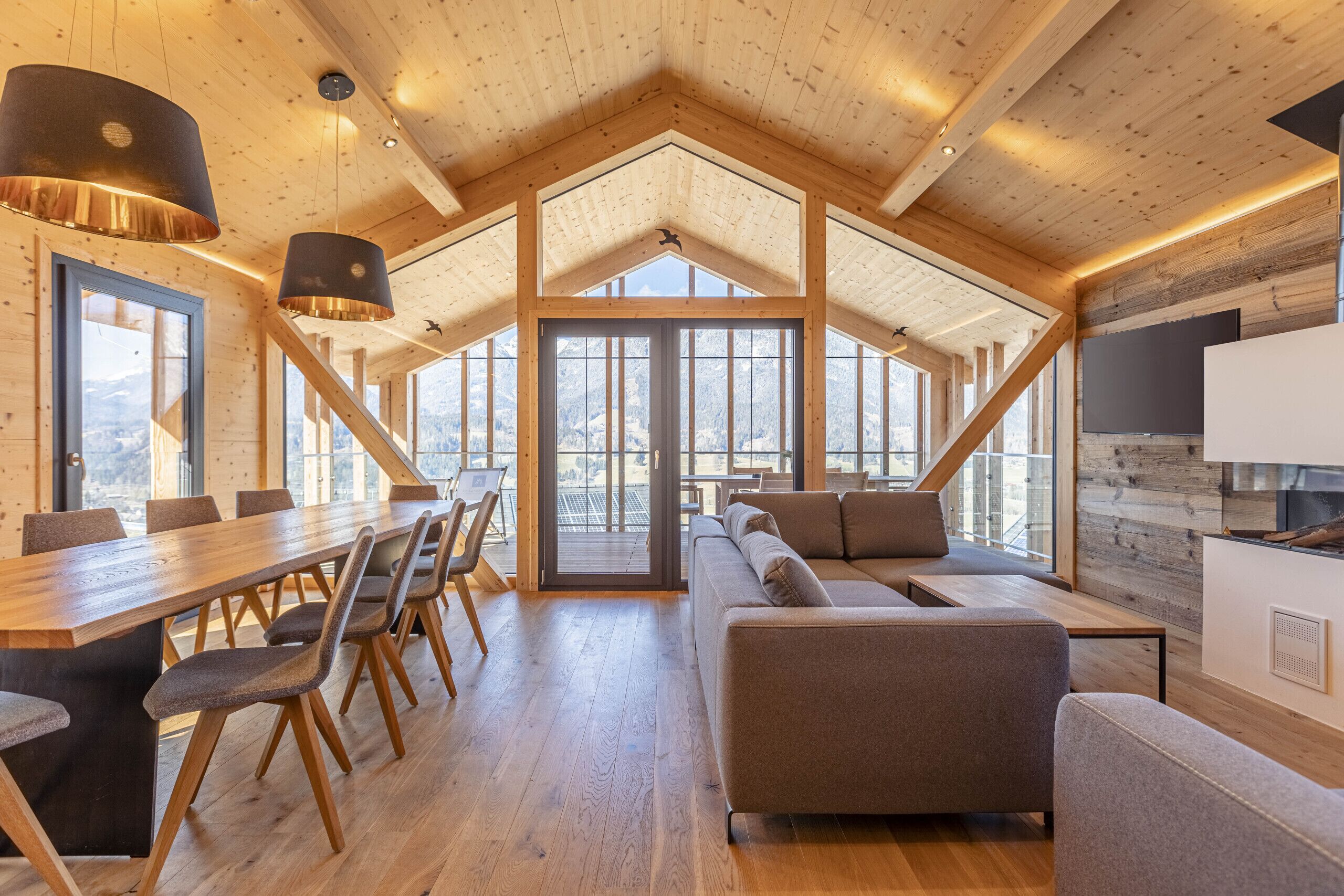 Ein geräumiges Zimmer mit hohen Decken, Holzbalken, einem langen Esstisch mit Stühlen, einer Couch und einem an der Wand befestigten Fernseher.