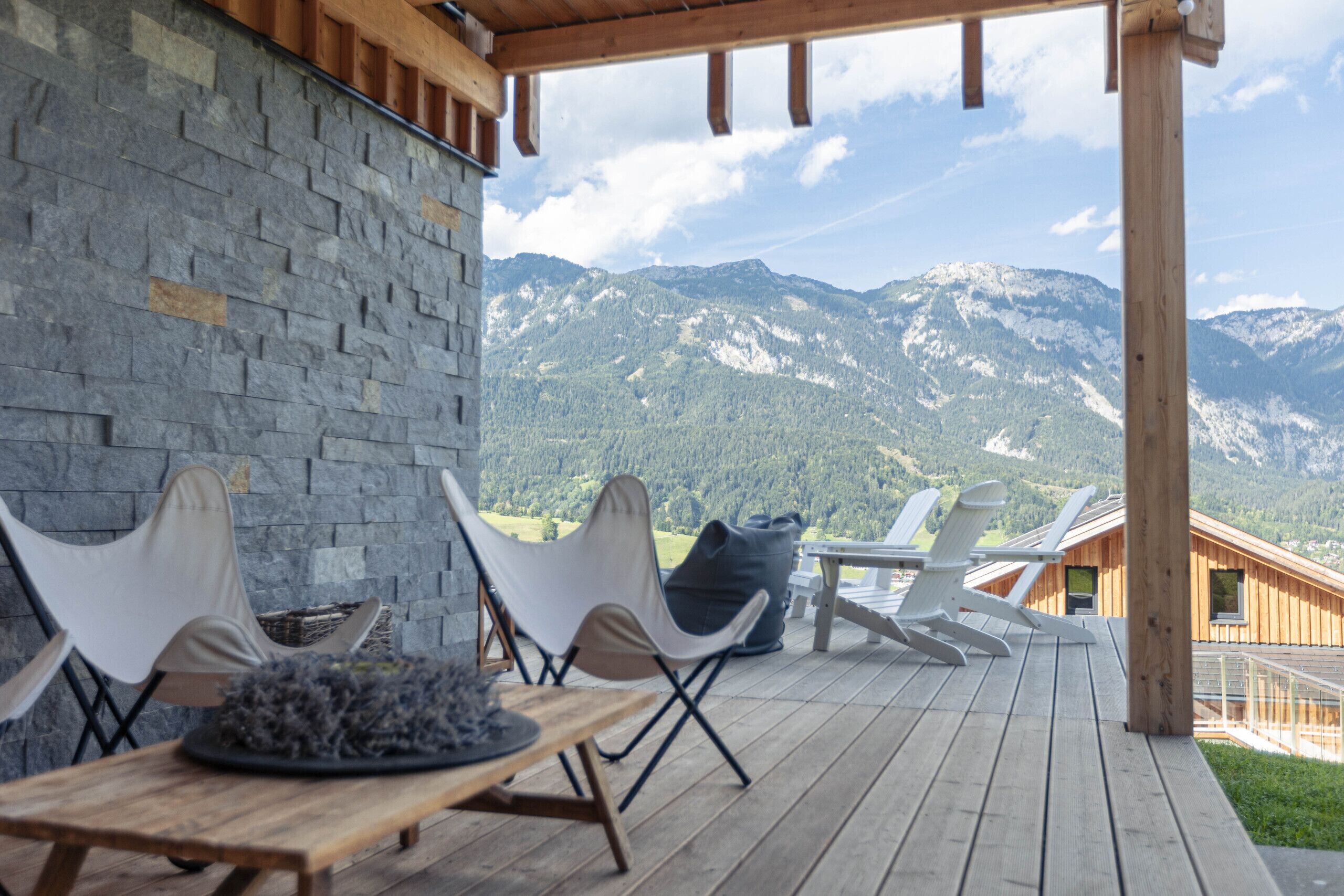 Eine Holzterrasse mit einer Steinsäule, Sitzgelegenheiten im Freien und Blick auf die Berge.