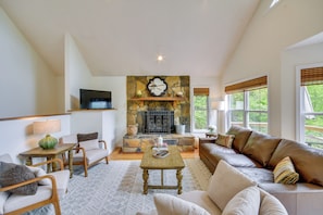 A living room with a stone fireplace, a sofa, a television, and a window with blinds. - Mountain Views & Hot Tub - Your Blue Ridge Getaway (Weaverville)