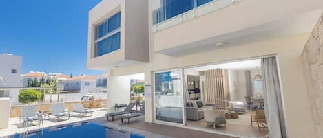 Un edificio moderno con piscina, sillas de descanso y una puerta de vidrio que da a un espacio interior con mobiliario.
