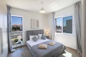 Ein Hotelzimmer mit Bett, Nachttisch mit Lampe, Fenster mit Vorhängen und Blick auf Gebäude.