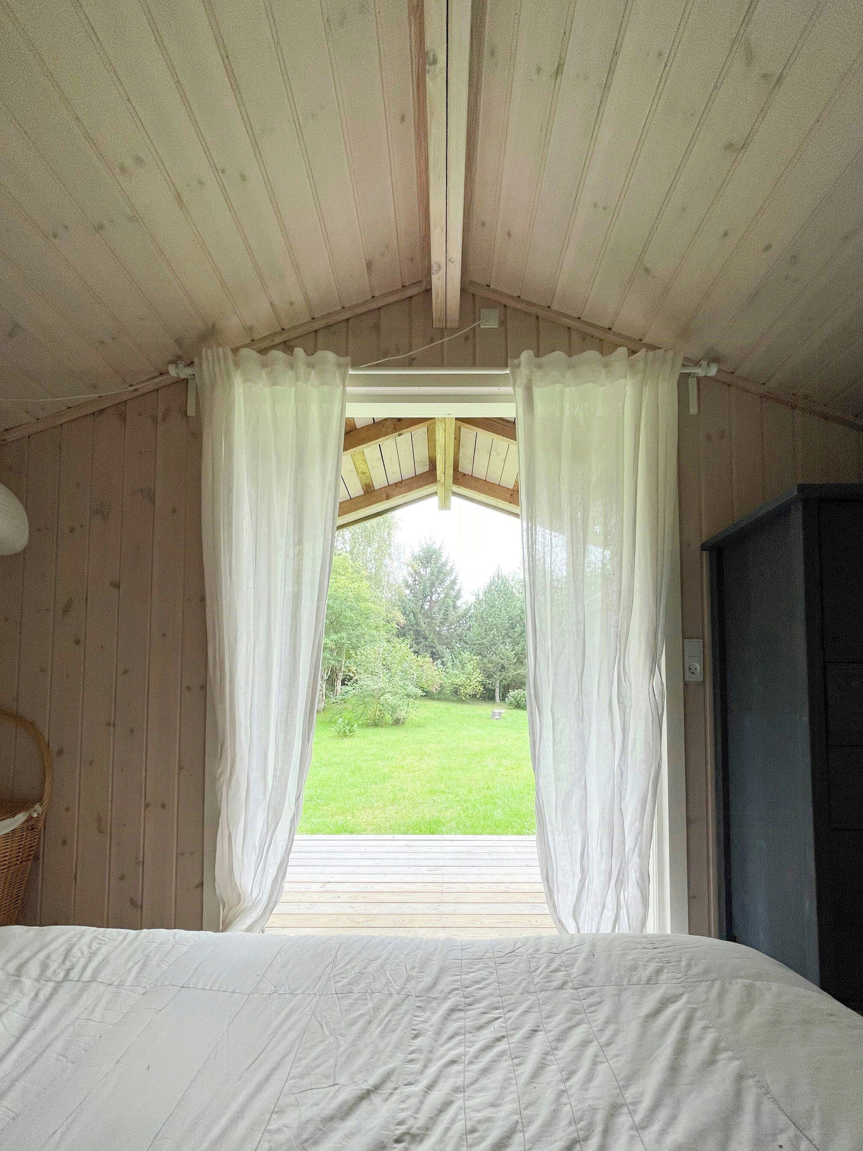 A room with a bed, wooden walls, a window with curtains, and a view of a green landscape.
