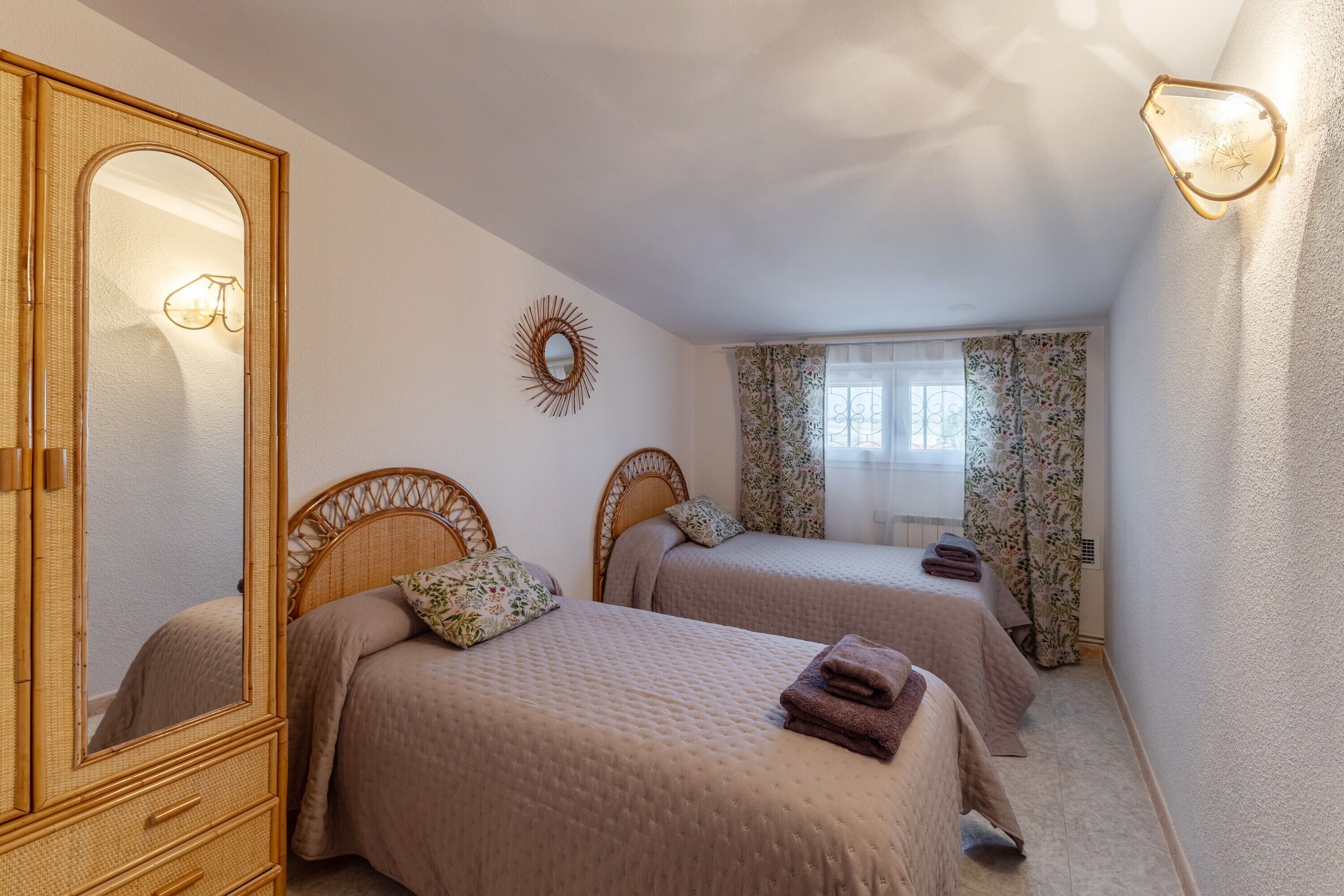 A bedroom with two beds, a large mirror, a wall-mounted light fixture, and a window with floral curtains.