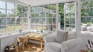 Un invernadero con un sofá blanco, una mesa de madera y vistas al exterior con vegetación.