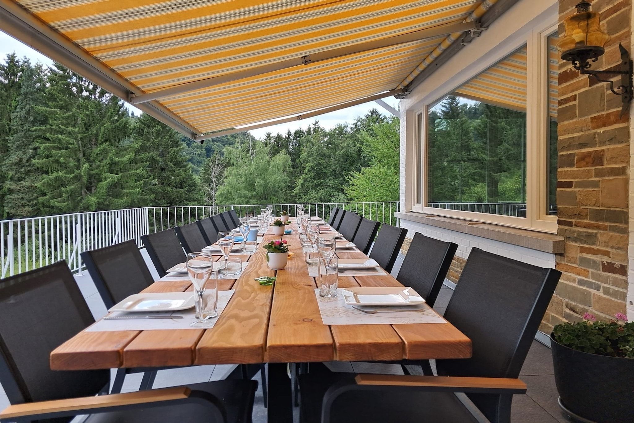 An outdoor dining area with a long table set for a meal, surrounded by chairs, under a striped awning, with a view of trees and a brick wall.