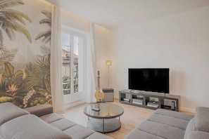 Ein Wohnzimmer mit rundem Glastisch, grauen Sofas, einem Fernseher auf einem Standfuß, einer Gitarre und einer Lampe.