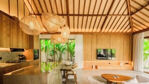 Un interior moderno con cocina, comedor y sala de estar, destacando vigas de madera, un televisor y un amplio ventanal con vegetación.