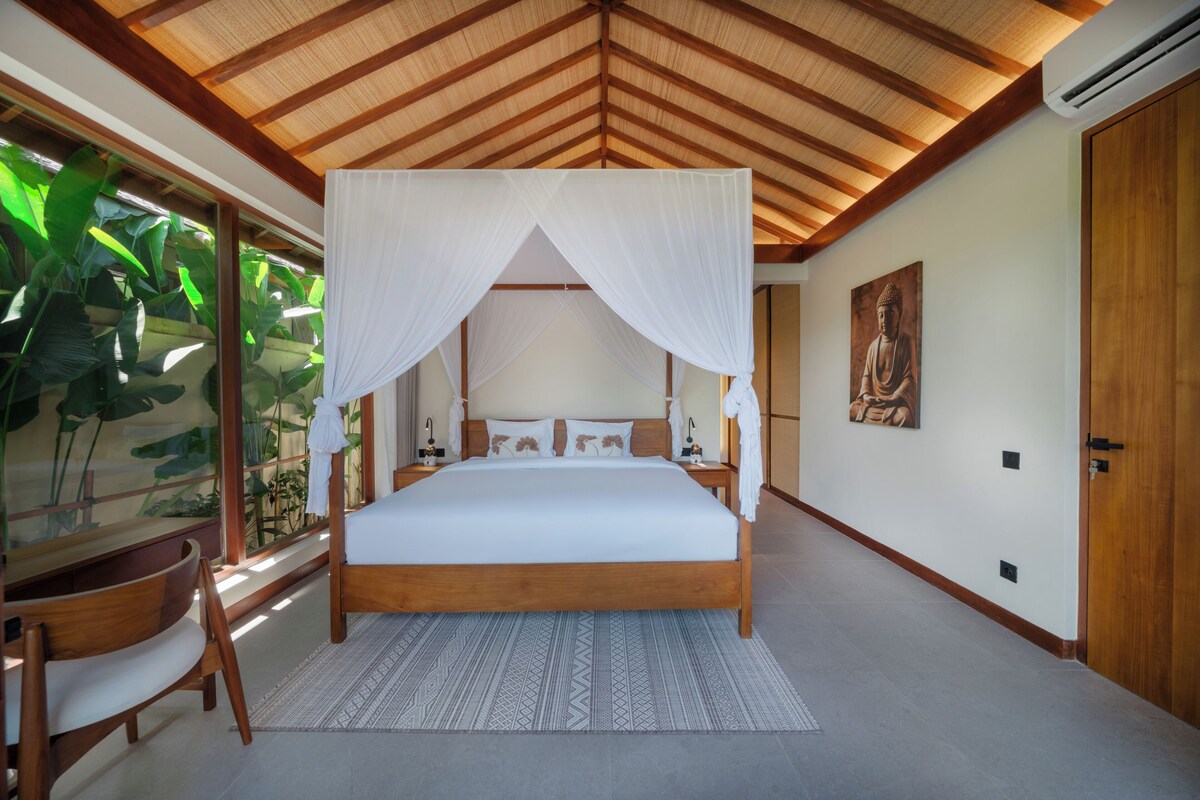 A spacious bedroom with a large bed, a canopy, a wooden ceiling, a large window with plants, a door, and a wall-mounted artwork.