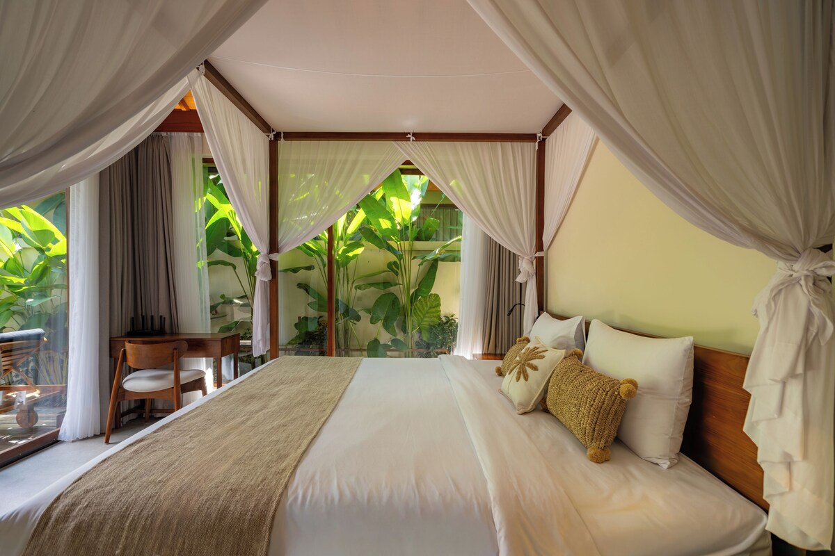 A bedroom with a canopy bed, a desk, and chairs, with a view of lush greenery outside.