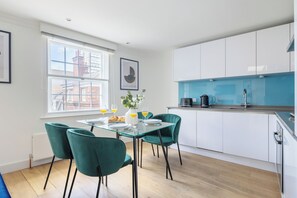 A modern kitchen with white cabinets, a glass dining table, and teal chairs. - Stylish Soho 1BR Central (London)